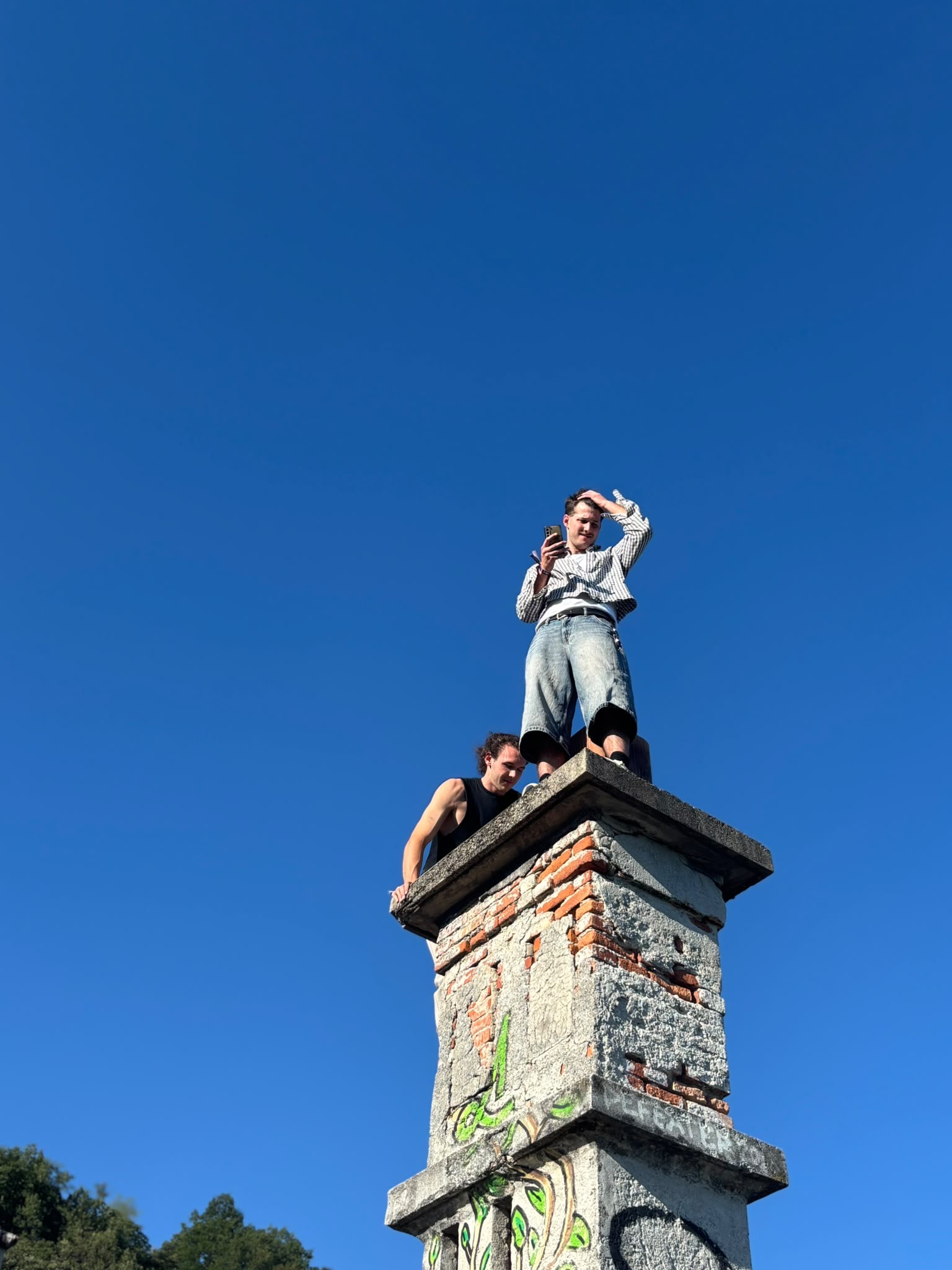 Zwei stehen oben auf einem alten Betonpfeiler vor tiefblauem Himmel, einer filmt mit dem Handy.