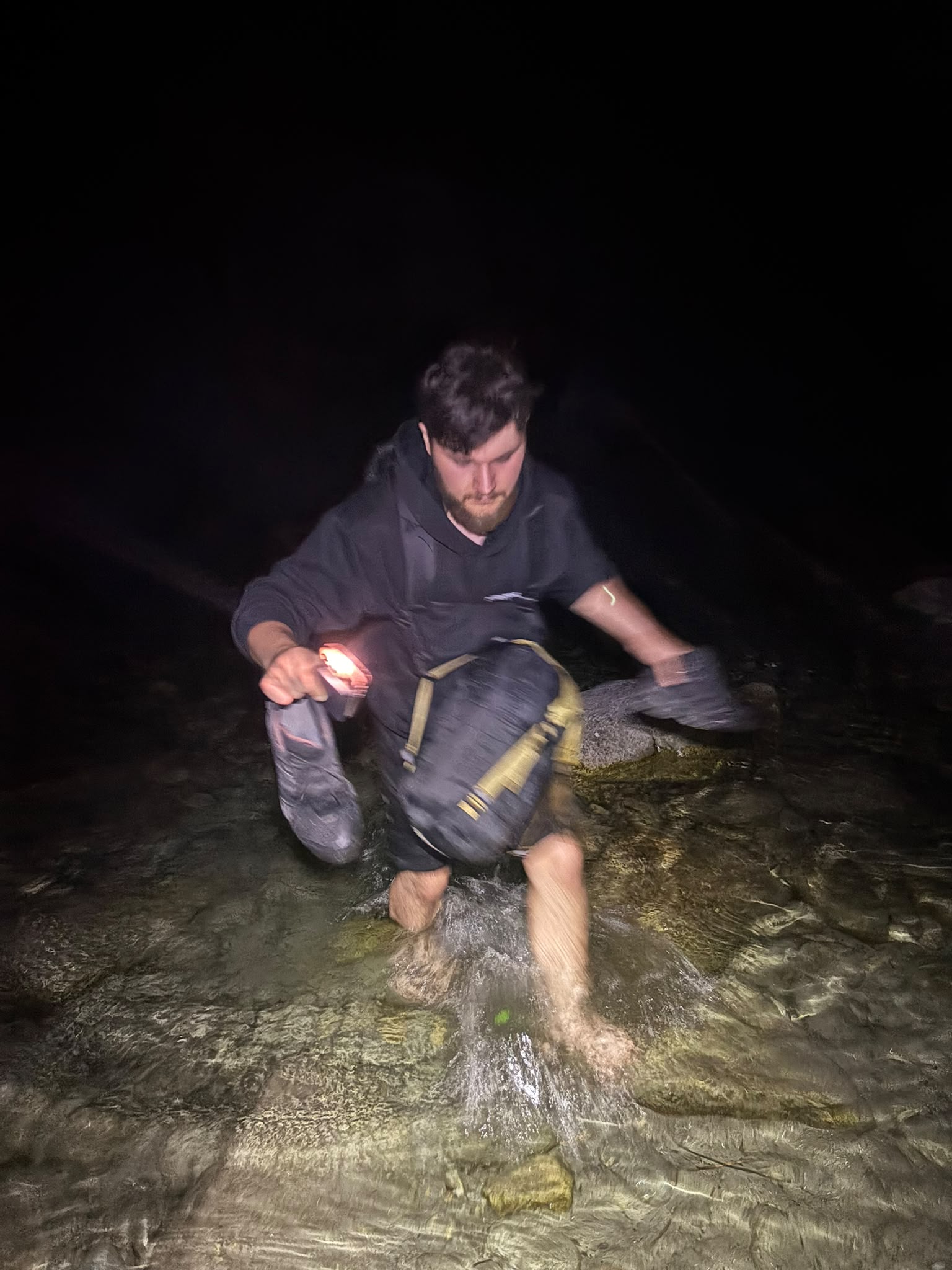 Nachts im Fluss: einer watet mit Rucksack, Schuhen und Lampe in der Hand durchs Wasser, vom Blitz verwackelt.