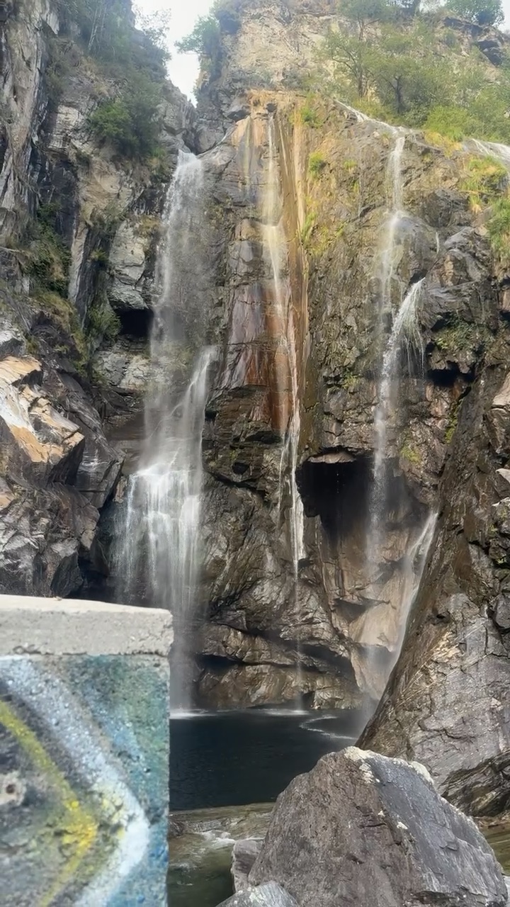 Betonmauer mit Graffiti als Absprung, dahinter stürzt ein Wasserfall über dunklen Fels in ein Becken.