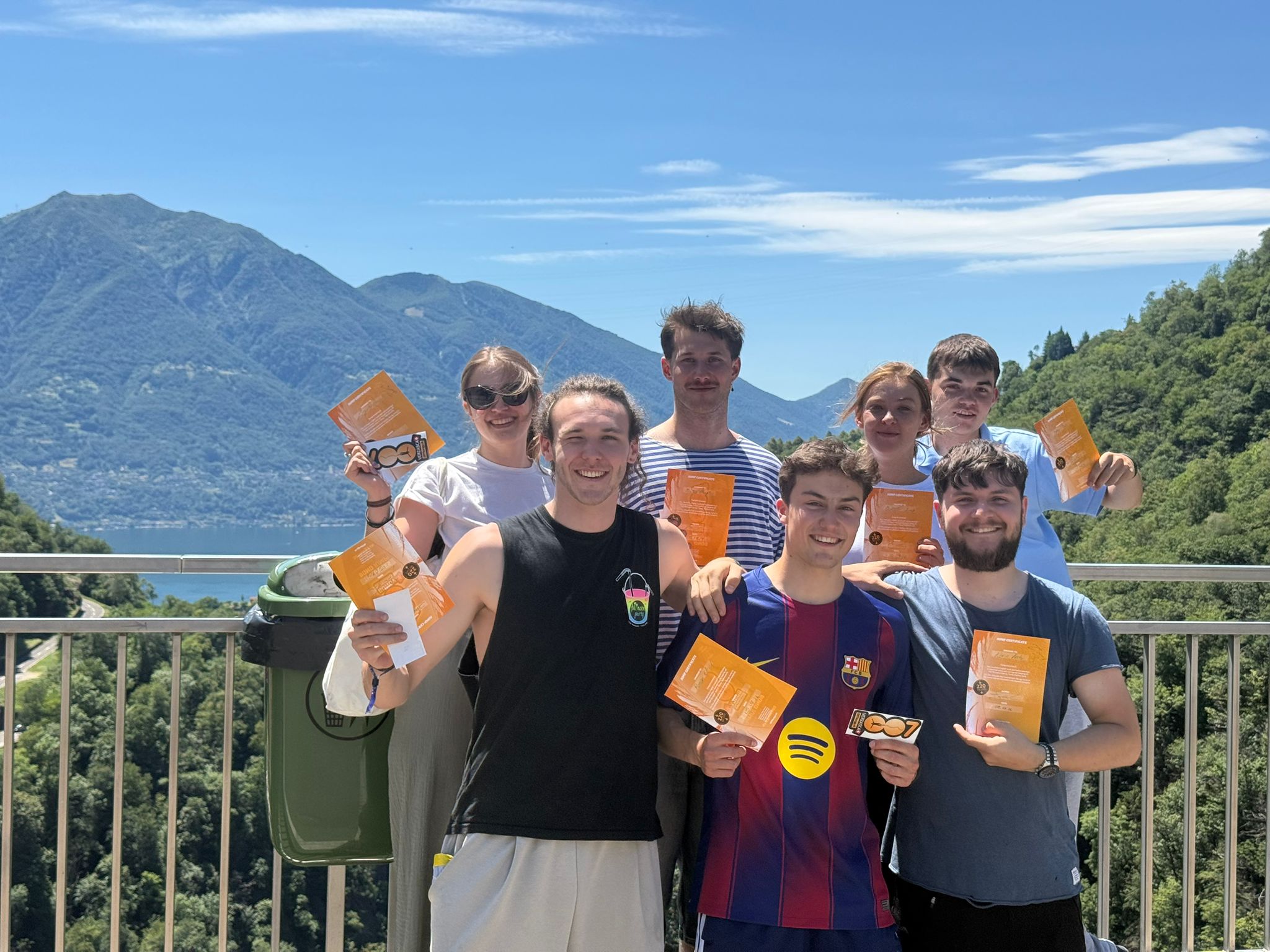 Sieben Leute lachen in die Kamera und halten ihre Sprungzertifikate hoch, hinter ihnen Lago Maggiore und Berge. Vorne von links: Jonas, Ramon und Kevin.