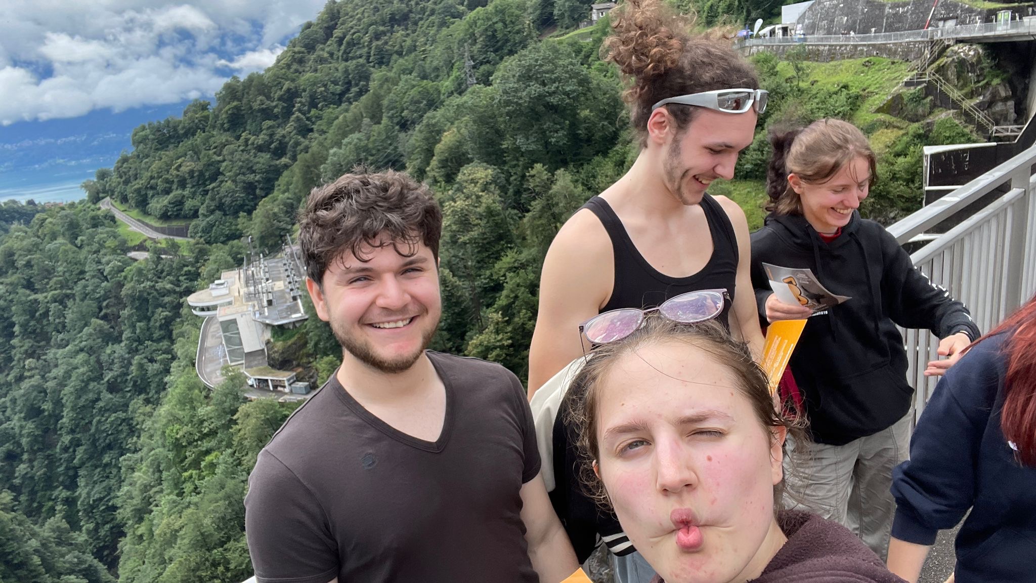 Gruppenselfie oben auf der Staumauer, alle aufgekratzt kurz vor dem Sprung, hinter dem Geländer fällt das Tal ab.